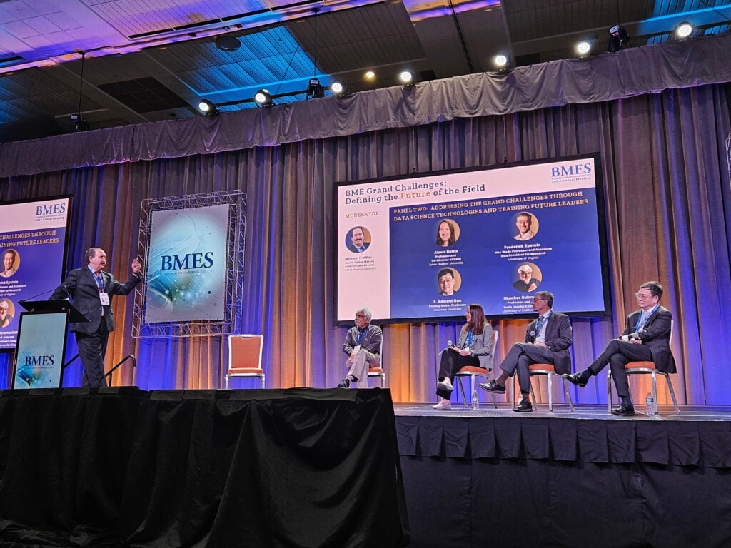 A man stands at a podium on stage with four other people sitting in chairs.